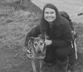 Greyscale image of a white female with long dark hair by the coast with her Dutch cross dog