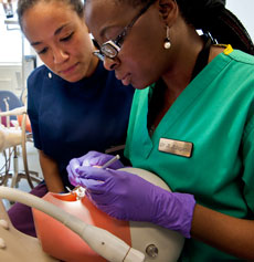 Female dental tutors in foreground wearing green dental scrubs demonstrating dental procedure on phantom head to female student in the background