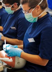 Female Student in foreground wearing dental scrubs and blue gloves performing procedure on phantom head with male student in background