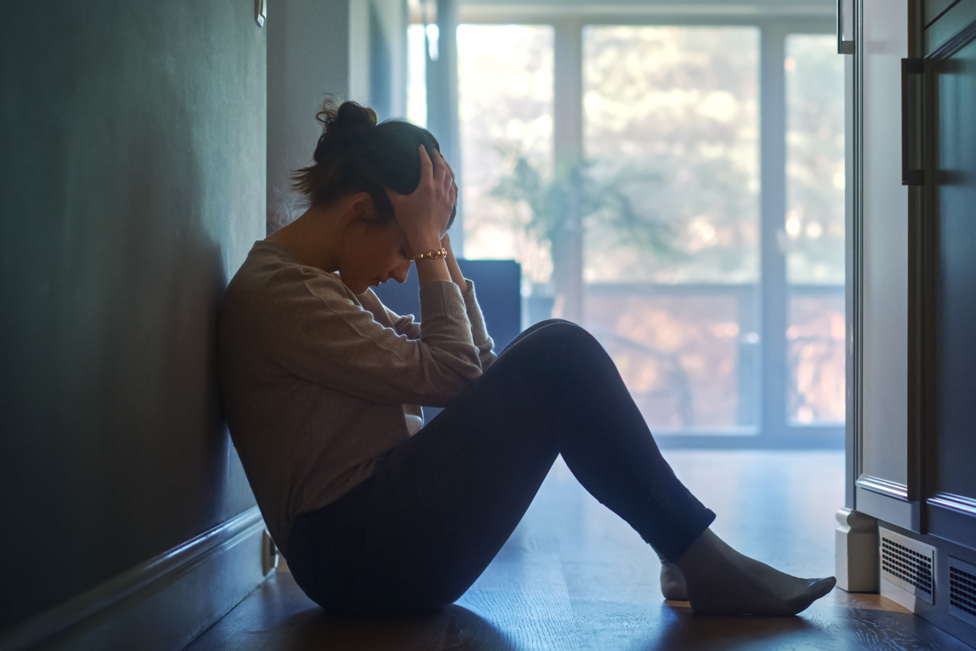 Sad young woman sitting on the floor in the hallway of her apartment, covering face with hands.