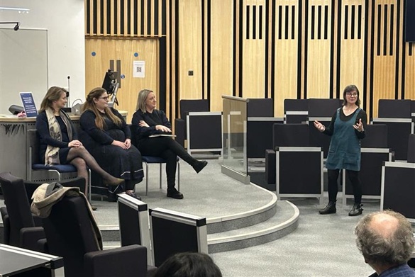 Professor Jo Meehan standing in front of attendees talking to a panel of speakers