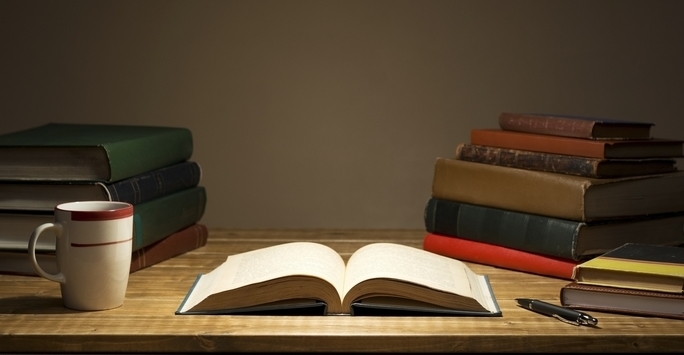 An open book on a desk under lamplight, coffee mug and stacks of books also on the table