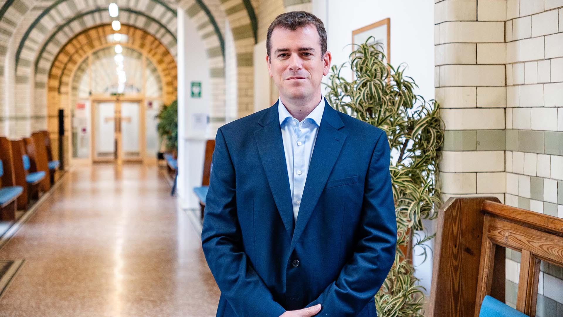 A wide 2 shot of the Dean of Medicine, Prof Andy McKeown. He is in a blue suit and has brown hair. Behind him is a tiled corridor in a medical building.