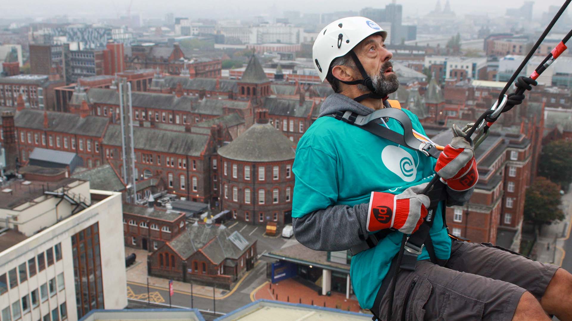 Justin Shanahan abseils down the side of a glass building. He is hung over the side, in the background the University campus unfolds behind him.