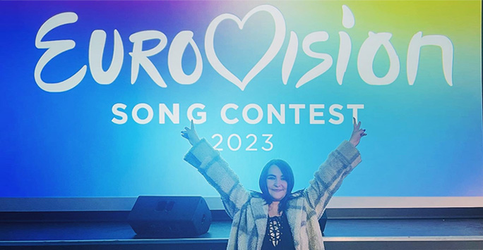A young woman stood in front of a multi-coloured Eurovision poster with her arms raised in celebration