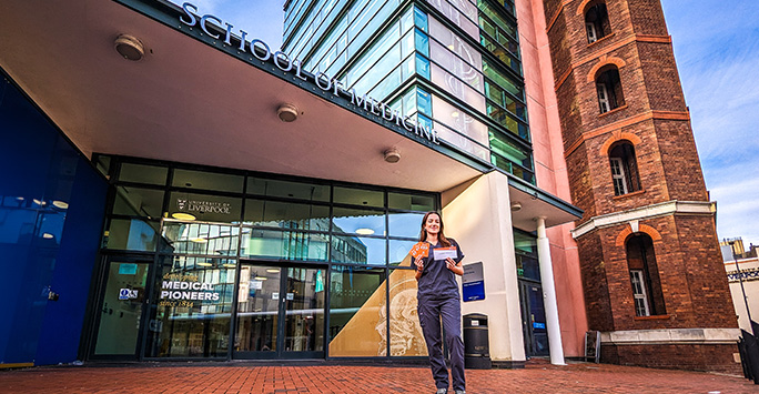 A young female student in scrubs, holding a small orange handful of paper outside of a modern office building, on which is the sign 