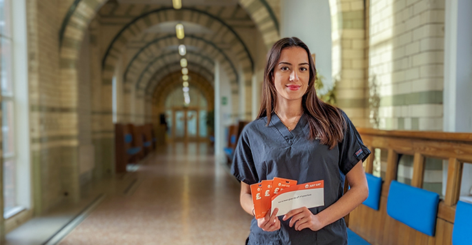 A young female student in scrubs, holding a small orange handful of paper in a long corridor. The text on the paper says 