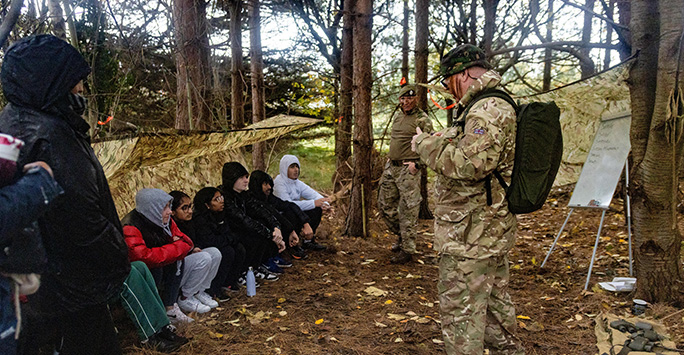 Students are sat under a tarpaulin in a wooded area, it is raining. In front of them stands an Army Infantryman who is briefing them about a task.