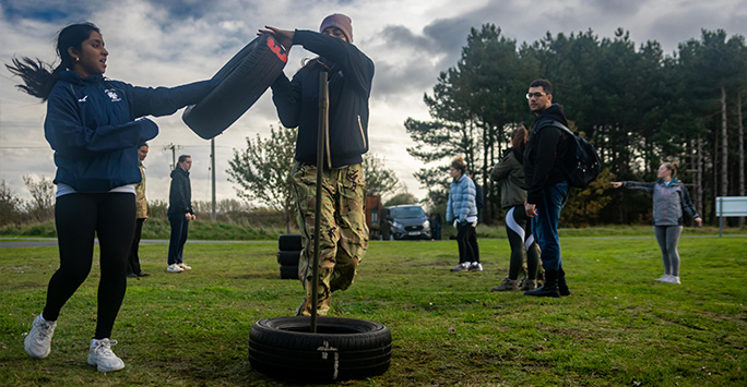 Two students are hoisting a car tire over a stand, whilst another directs others in the team in the background