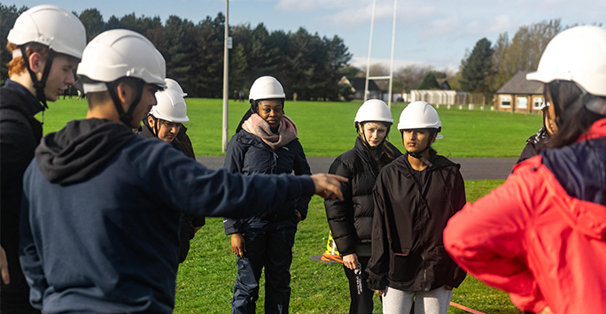 5 students conversing about a task, whilst wearing hard hats in an open field. One student is pointing out of frame to an aspect of the task to direct the other 4 students.