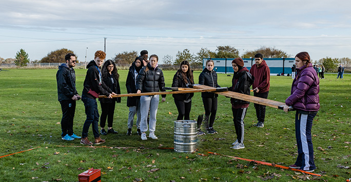 A group of students carrying 3 wooden planks made into a triangle formation as the solution to a task