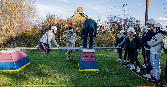 Students watching as their colleagues navigate a challenge of sliding over two metal bars positioned between two gym horses