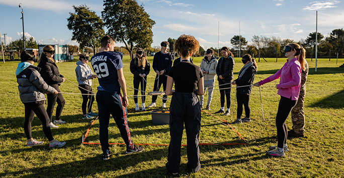 A group of students who are blindfolded being led in an exercise to pick up an object with a hook attached to a rope, whilst being directed by their unblindfolded colleague