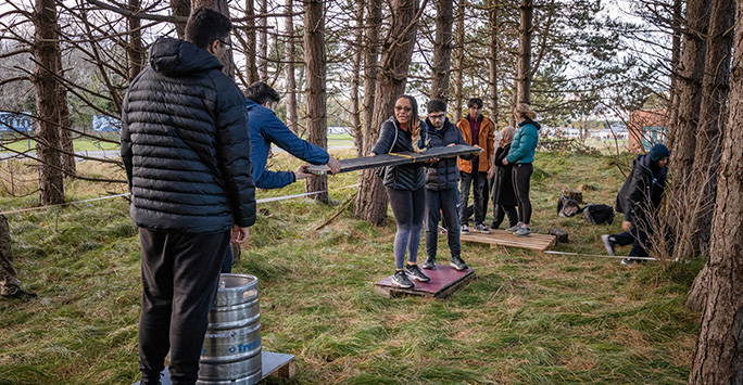 A group of students passing a wood plank along a line to form a river crossing