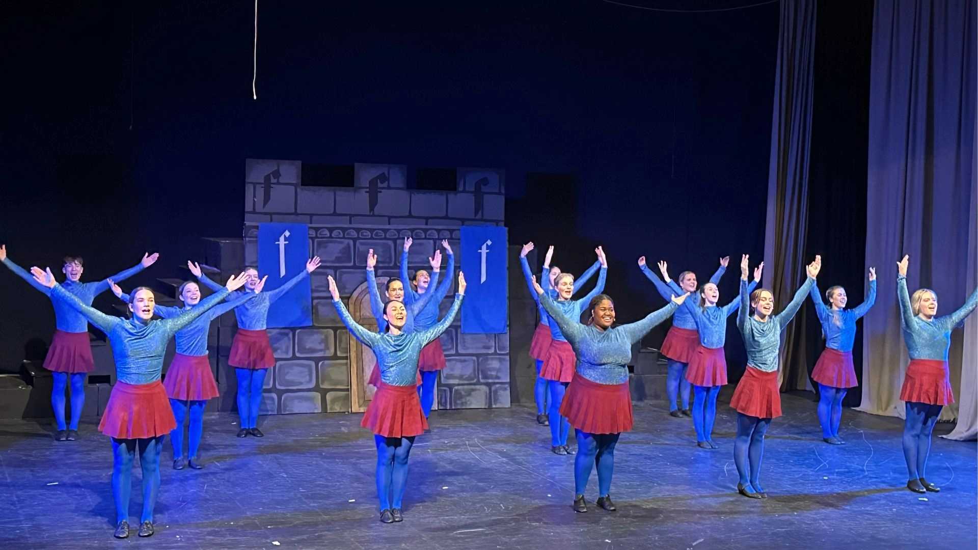 Ensemble cast performing a choreographed tap dance in blue and red costumes in front of a castle set.