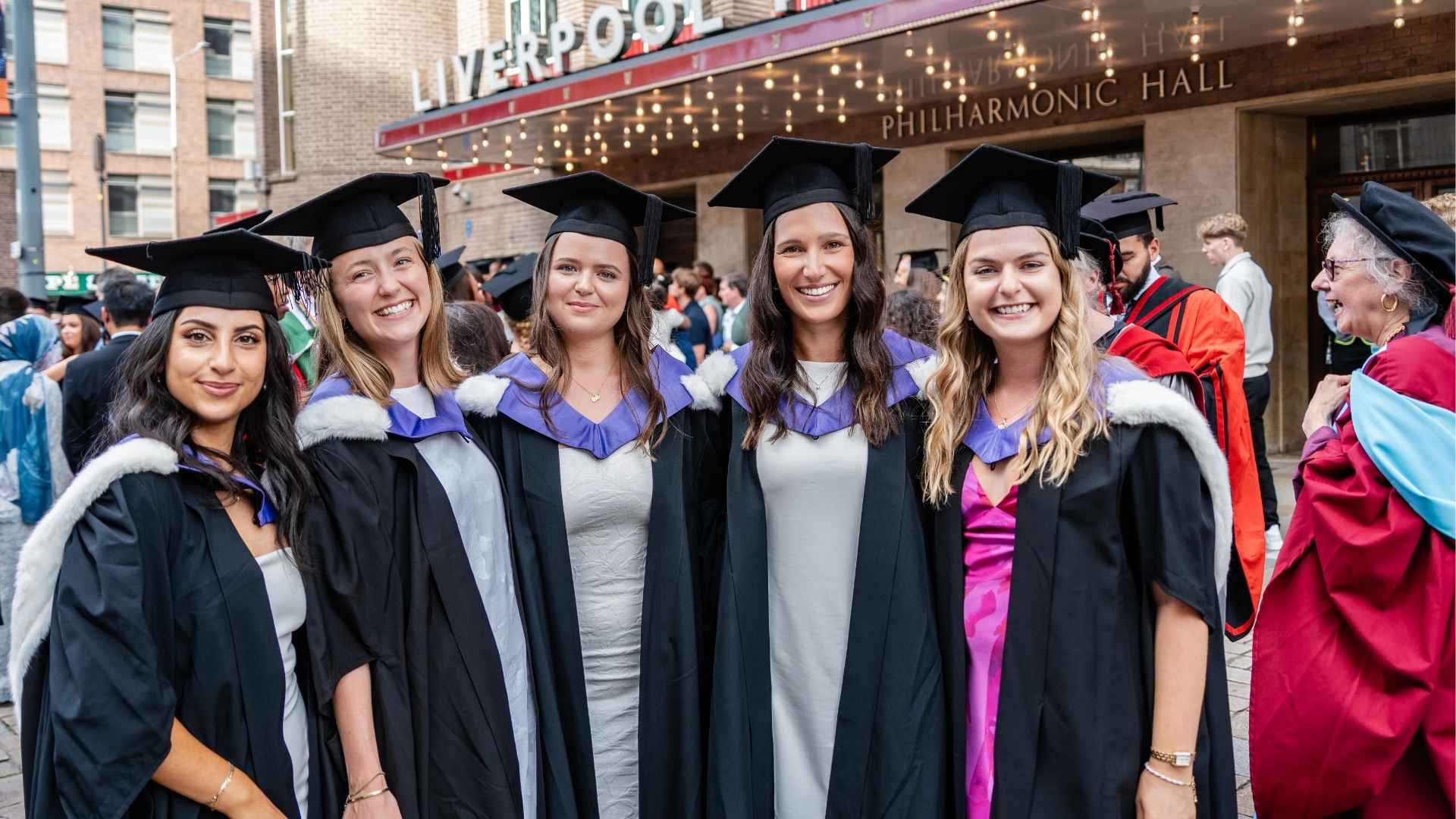 Five smiling graduates in caps and gowns pose outside the Philharmonic Hall, with other attendees in the background.