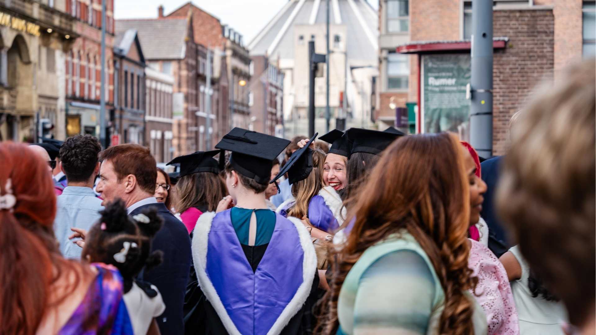 A graduate beaming as she hugs a friend in a crowd of fellow graduates, with the Metropolitan Cathedral visible in the distance.