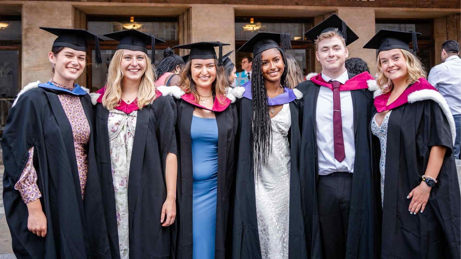 Six graduates in gowns and hoods smiling at the camera outside Liverpool Philharmonic Hall.