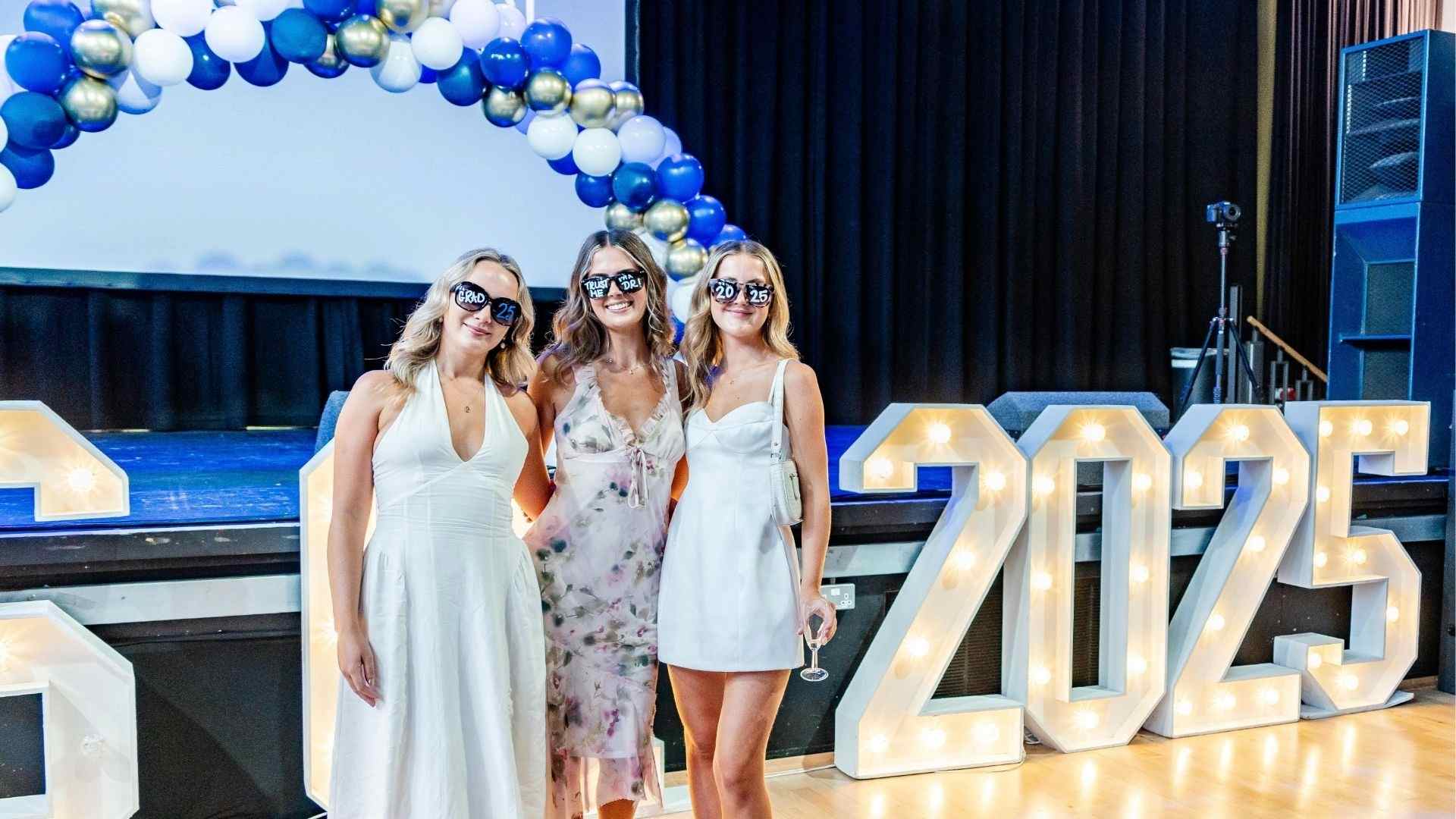 Three graduates pose indoors in front of a balloon arch and large lit 2025 sign, wearing celebratory glasses