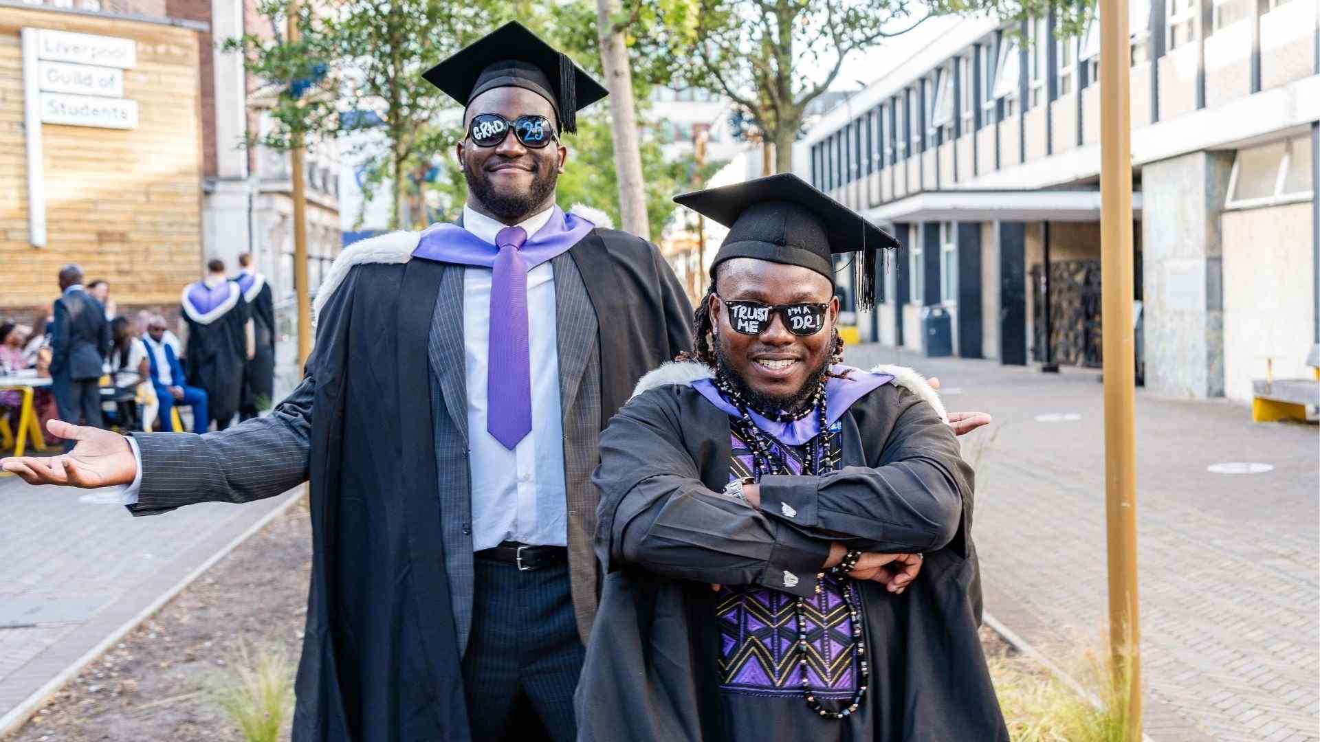 Two graduates in gowns and hoods smile proudly outside the Guild of Students