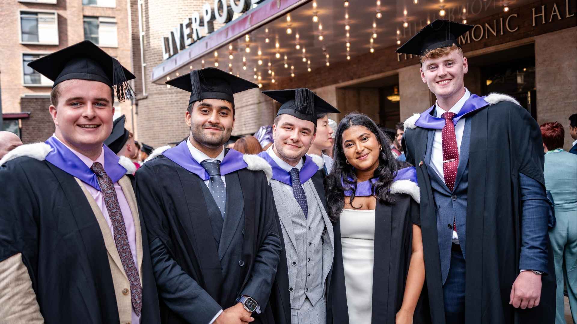Five graduates in caps and gowns pose outside Liverpool Philharmonic Hall, smiling proudly after the ceremony.