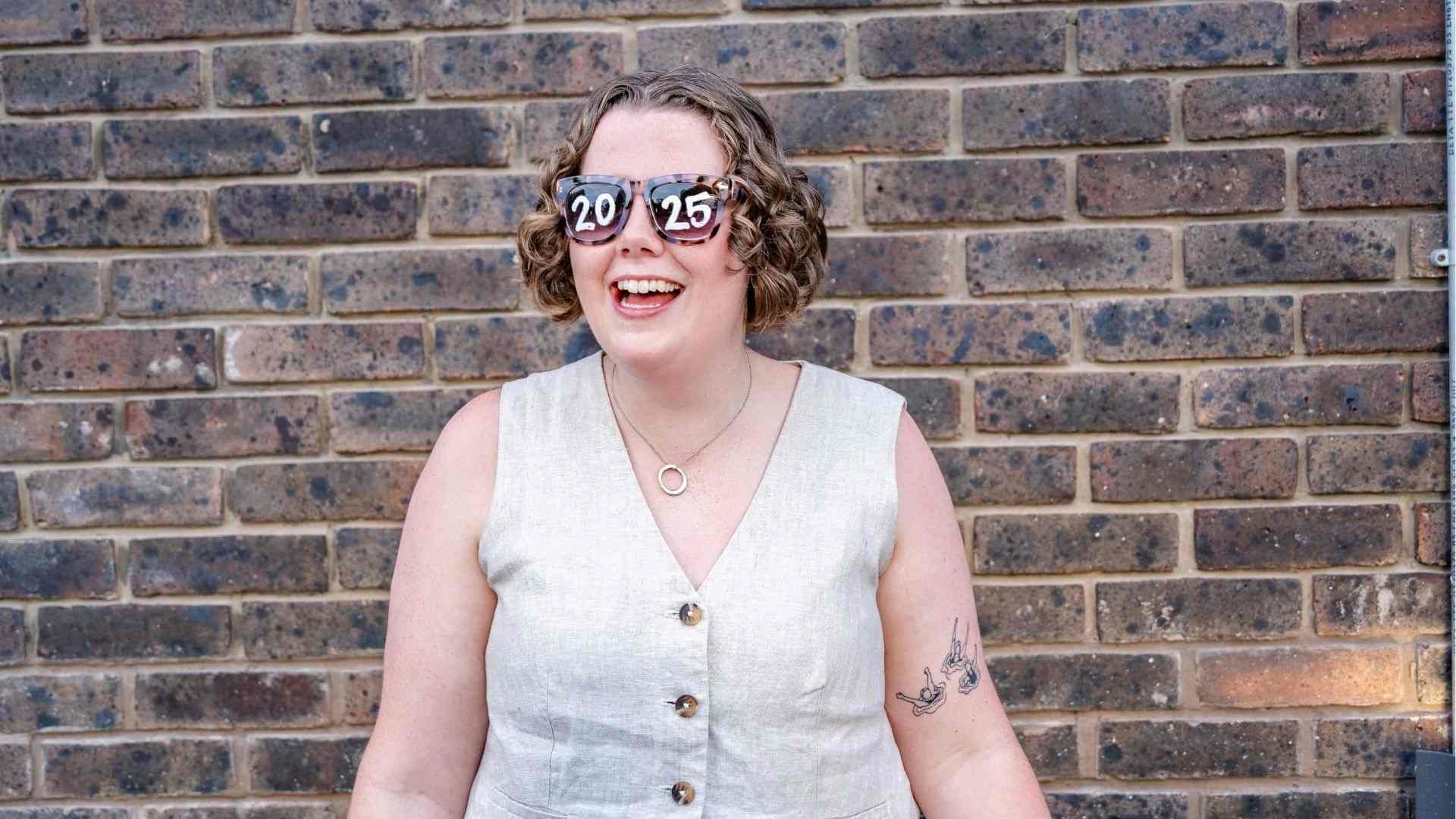 A graduate laughs while wearing oversized 2025 sunglasses, posing in front of a brick wall.