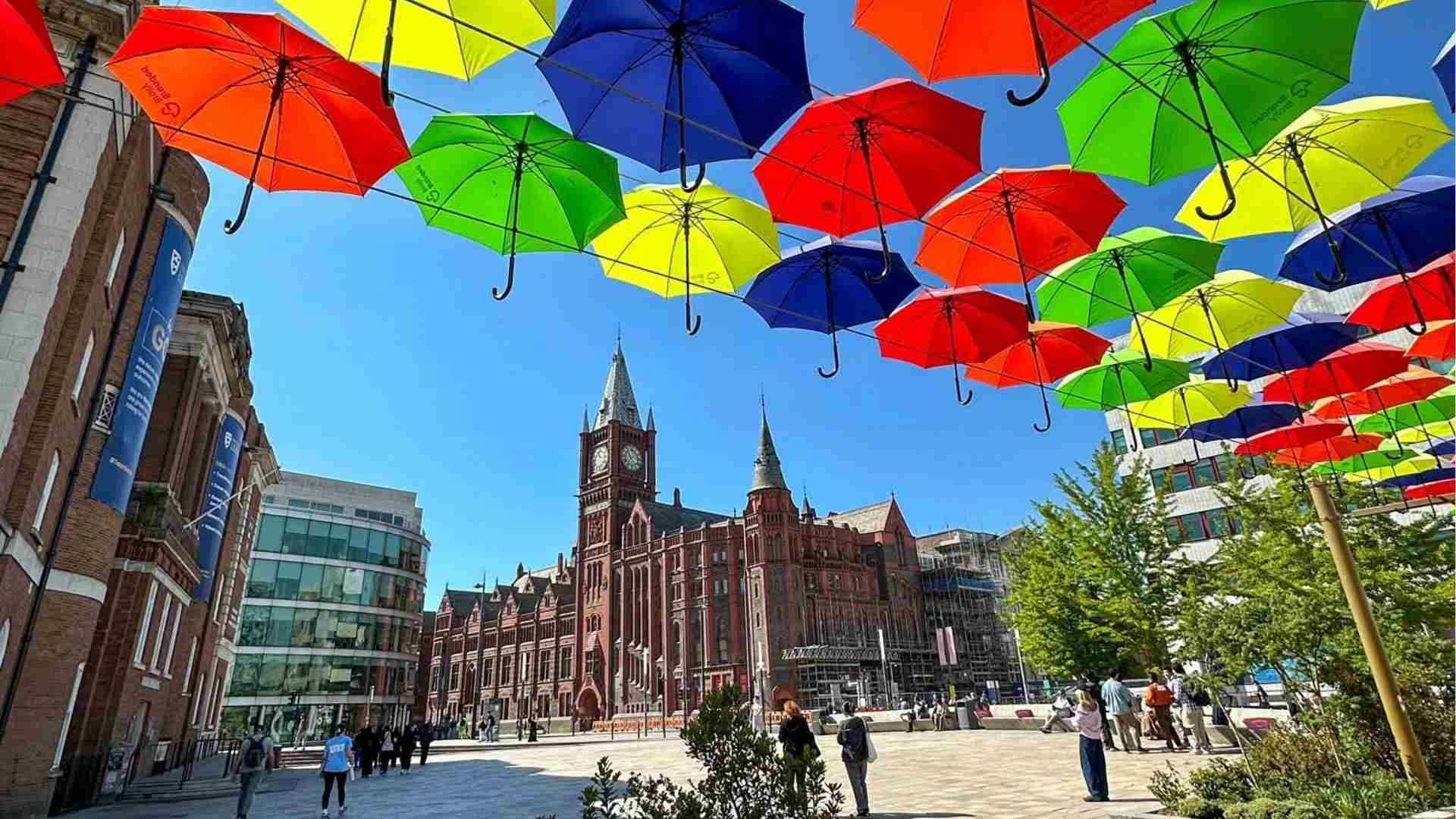 Colourful umbrellas suspended above University Square at the University of Liverpool, with the iconic red-brick Victoria Building and clock tower in the background on a sunny day