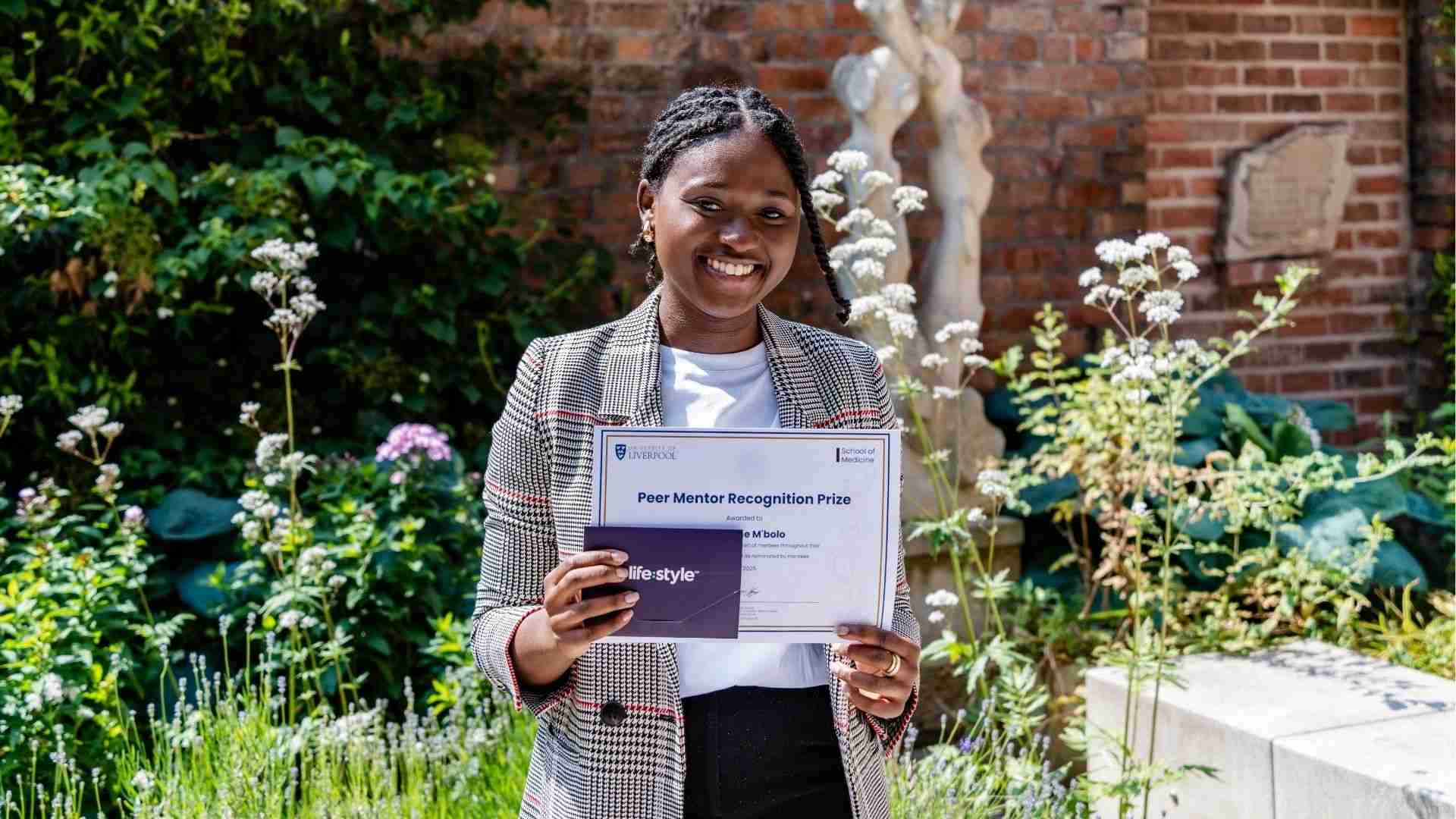 Ghislaine Mbolo smiling and holding her Peer Mentor Recognition Prize certificate and gift card, standing in a garden outside the University of Liverpool School of Medicine.