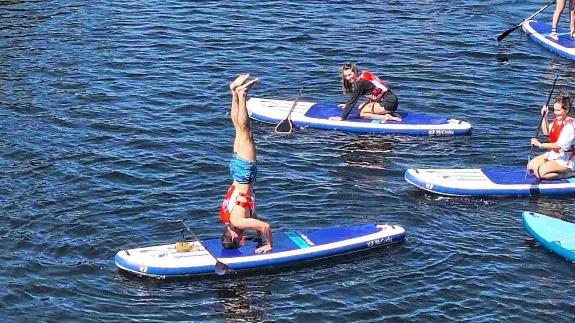 Student doing a headstand on a paddleboard while others watch and paddle nearby on the Liverpool docks.