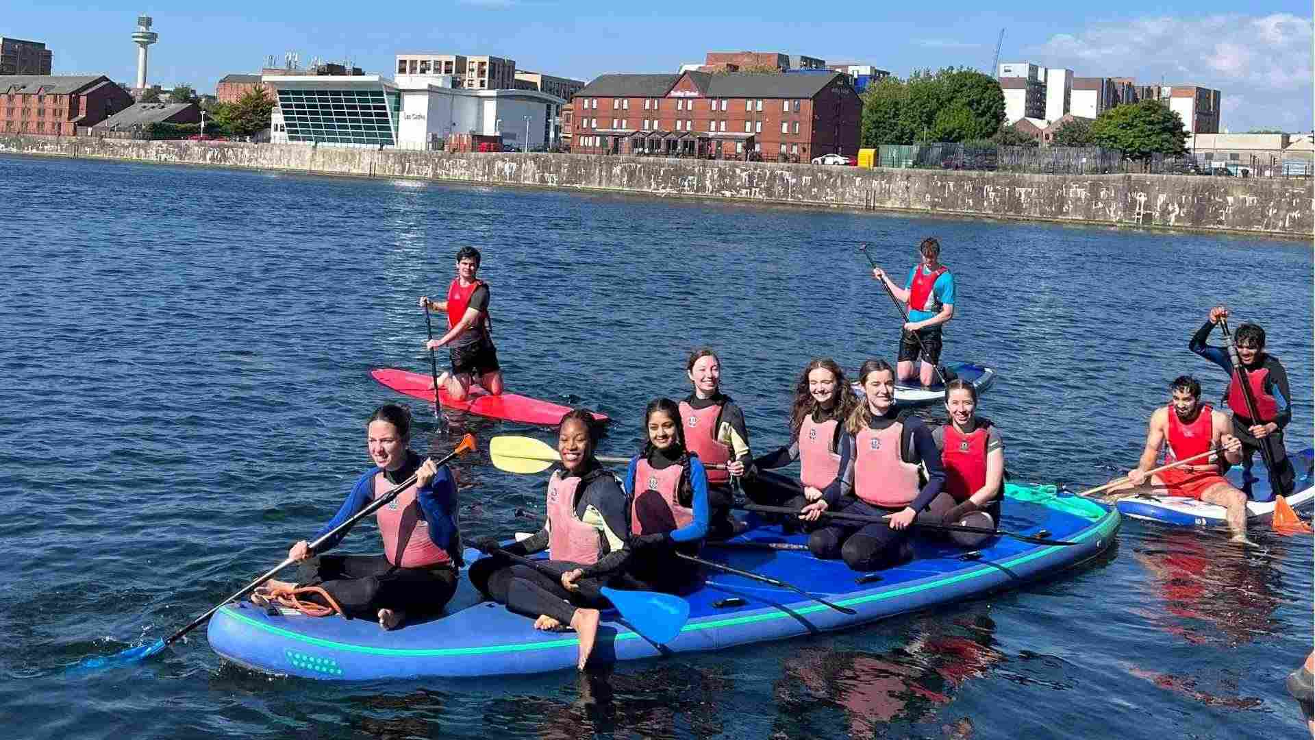 Group of students sitting on a giant paddleboard, smiling and enjoying the water.