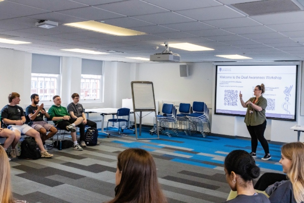 A facilitator introduces the Deaf Awareness Workshop to a group of student doctors sitting in a semi-circle. A presentation slide on screen welcomes attendees.