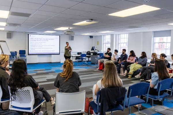 The facilitator delivers an engaging session to a wider group of student doctors, with an educational slide projected behind her.