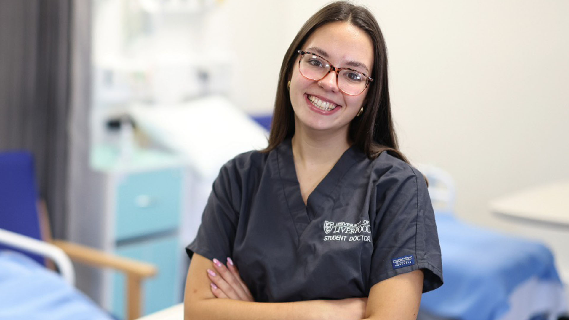 Woman student doctor in grey scrubs with arms folded standing in front of a hospital bed