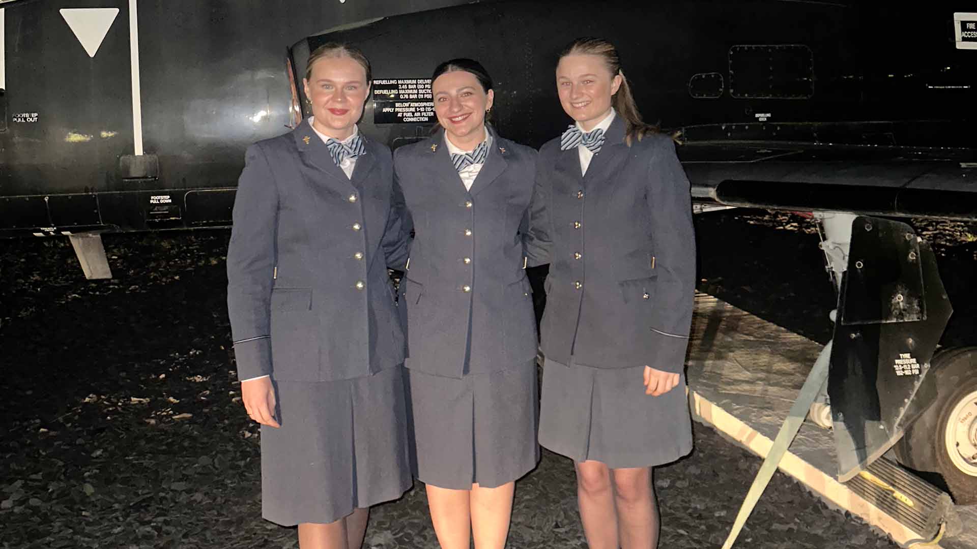 Three medical cadets in formalwear pose at night in front of a military aircraft
