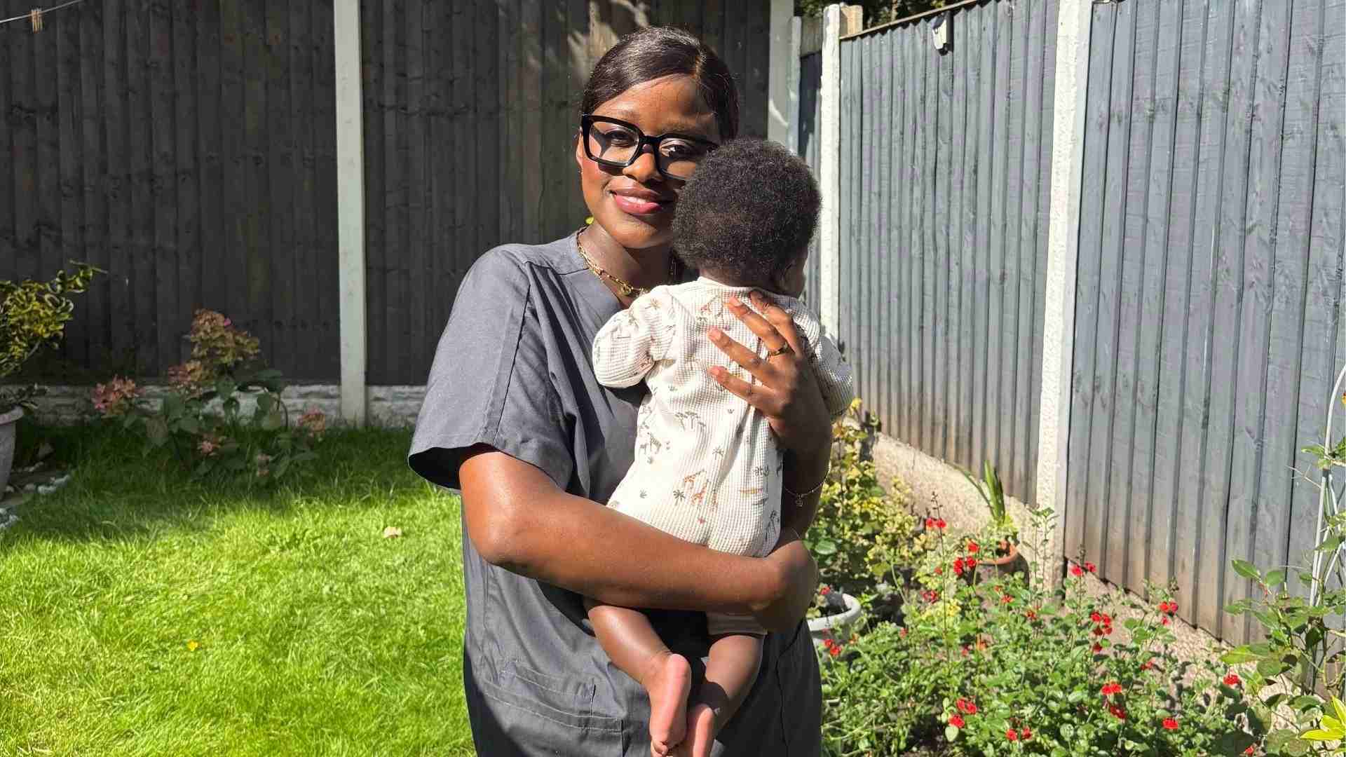 student doctor in grey scrubs cuddles her baby in sun kissed garden