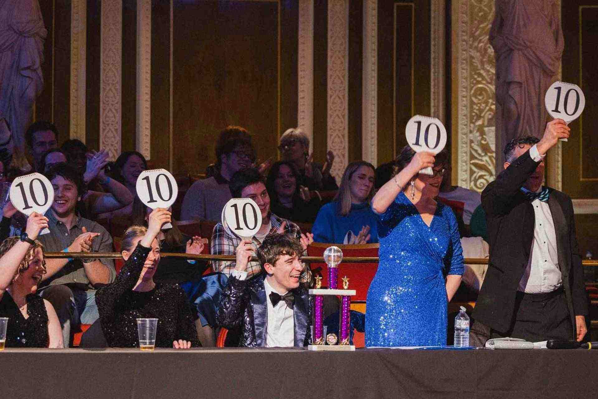 Professor Scott sits smiling and holding up a score card alongside other judges