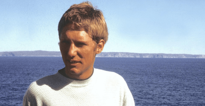 man with 1970s style haircut and white t shirt with blue sky and sea background