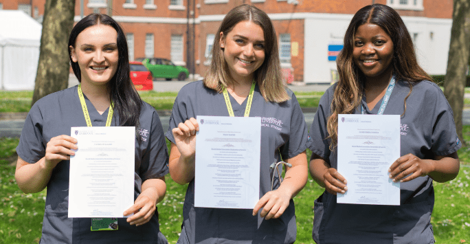 Three women in grey scrubs pose outdoors with certificates