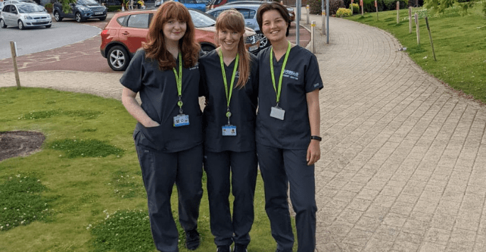 Three women in grey scrubs stand with their arms around each other outdoors, in front of a car park