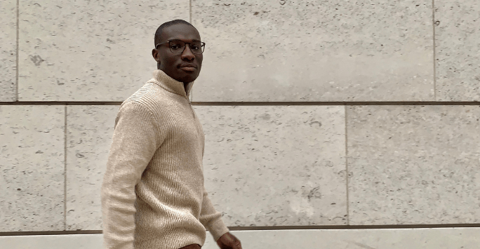 Man wearing glasses and a beige jumper walks to the right in front of a wall with large cream coloured bricks
