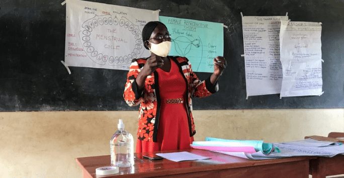 An educator stands behind a desk and in front of a chalkboard with diagrams about menstruation