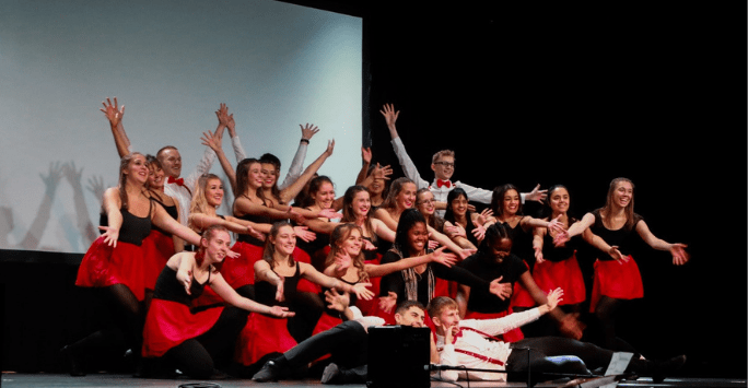 performers in black tops and red skirts raise arms and smile