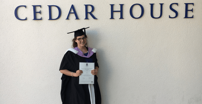 Graduate in cap and gown in front of Cedar House sign