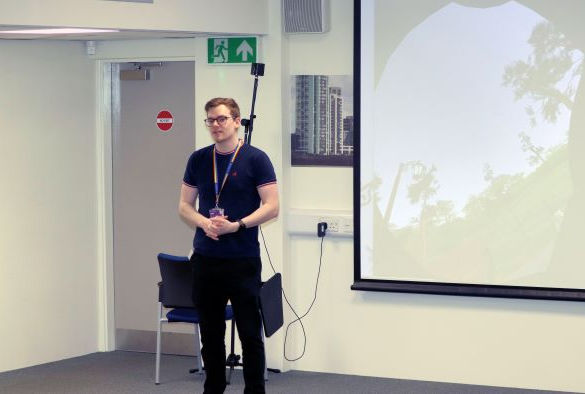 A lecturer stands in front a projection screen wearing a black tshirt and jeans