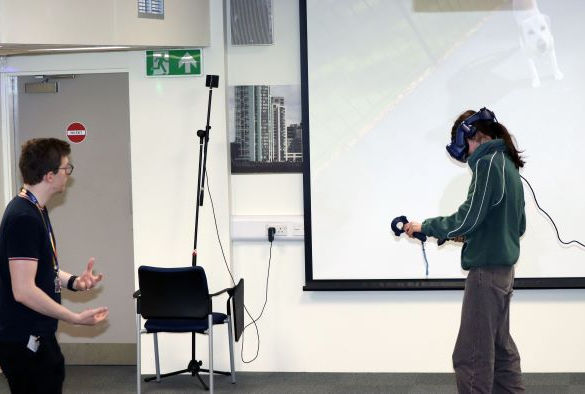 A student in a green hoody and joggers wears a VR headset in front of a screen