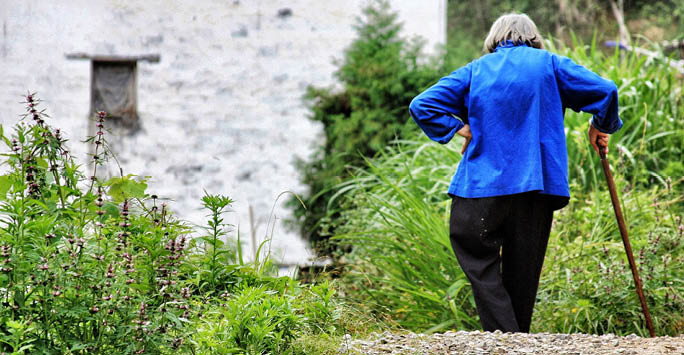 An older woman walking with her walking stick by a river.