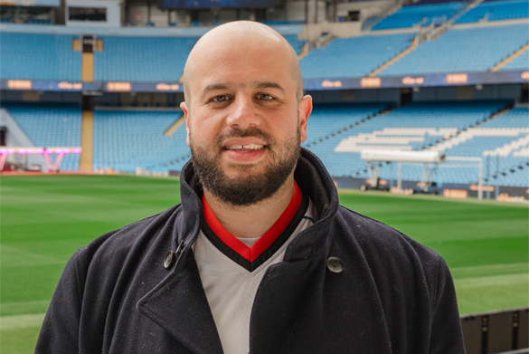 Dr Moosavi  stands inside a large football stadium, with green pitch and blue seating in the background.