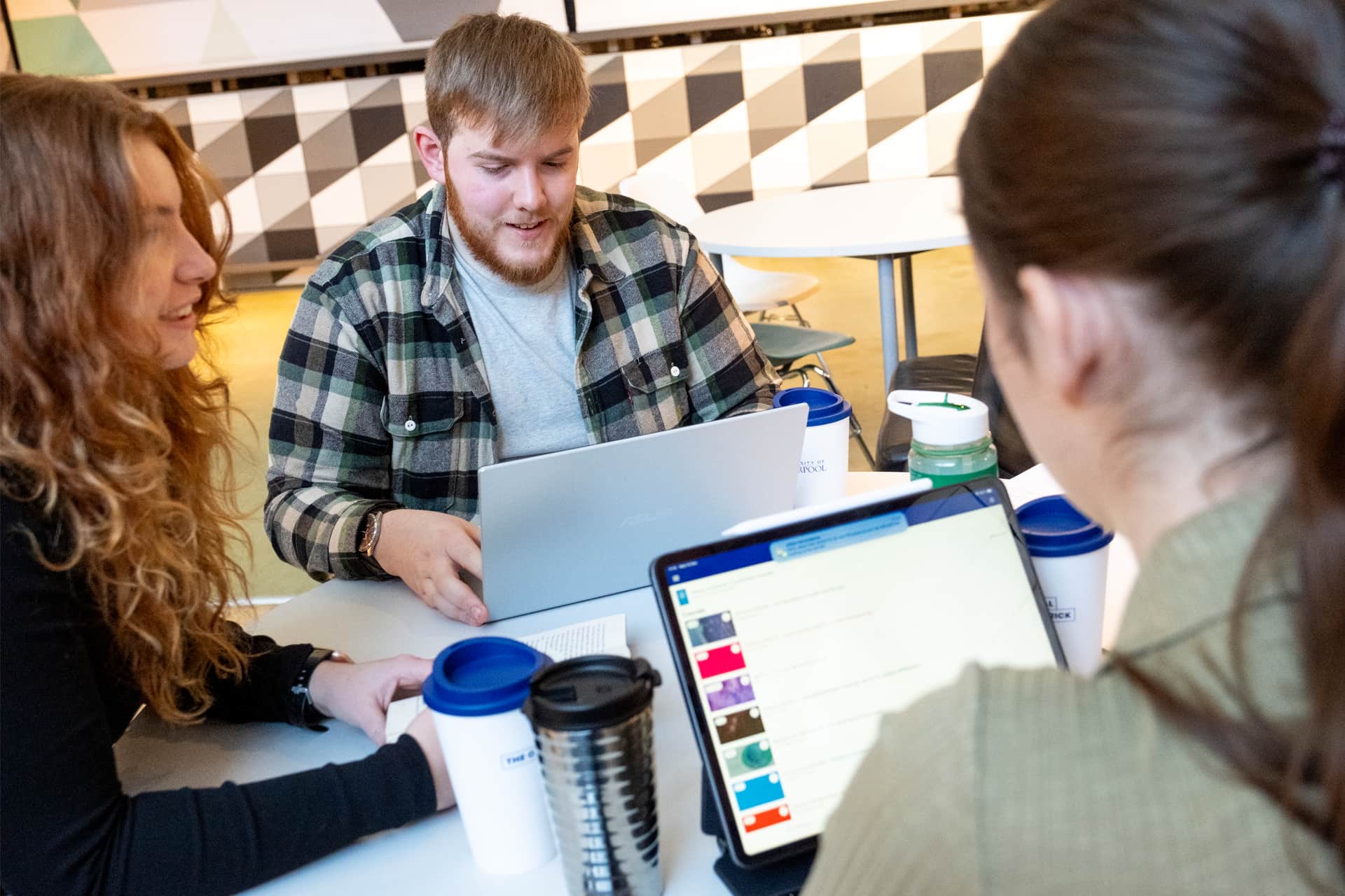 Three people sitting around a table in discussion, two using laptops.