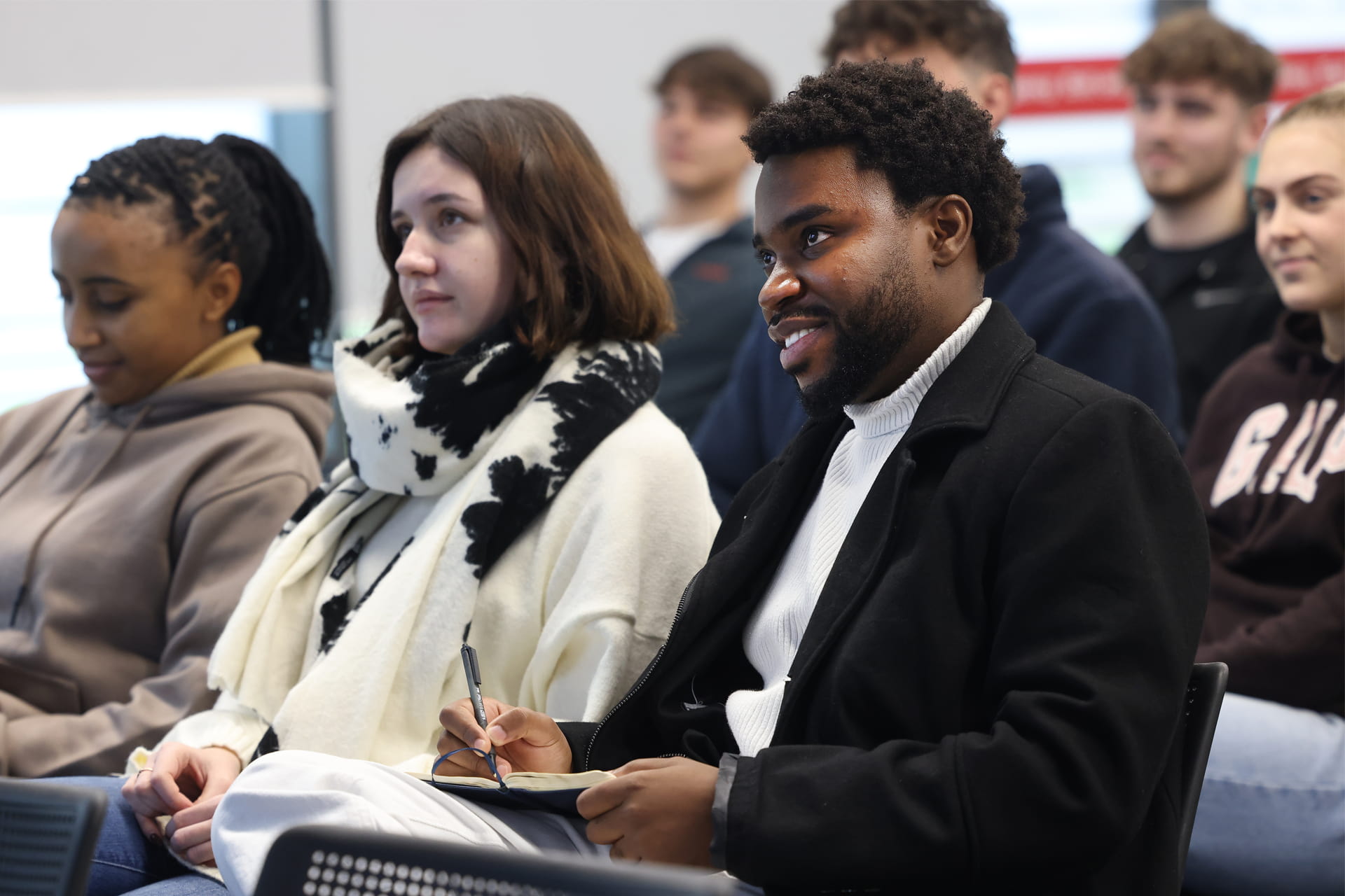 A group of student eople seated in a lecture setting, with three individuals in the front row attentively listening.