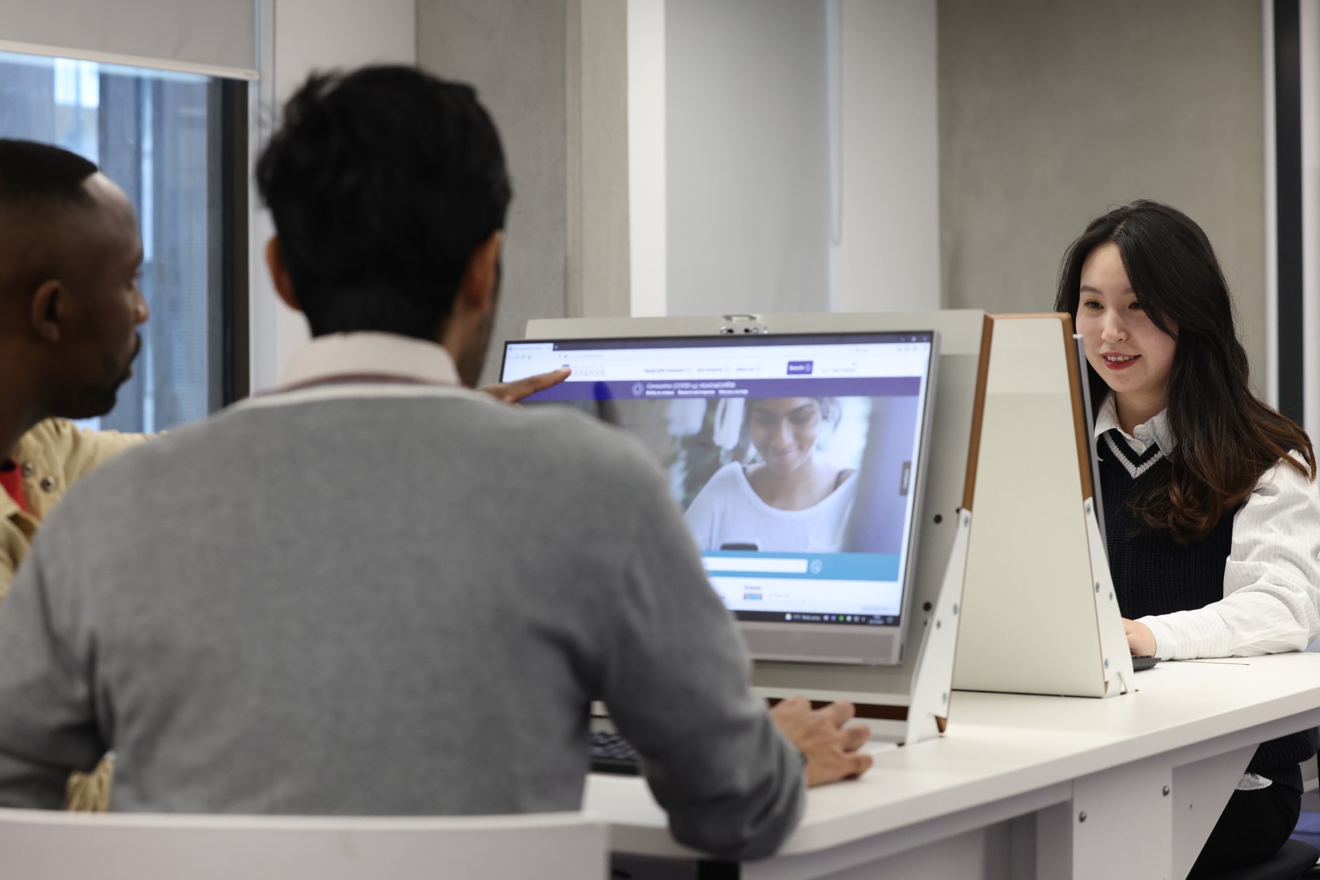 A group of students working together on a computer, with one joining them via video call.